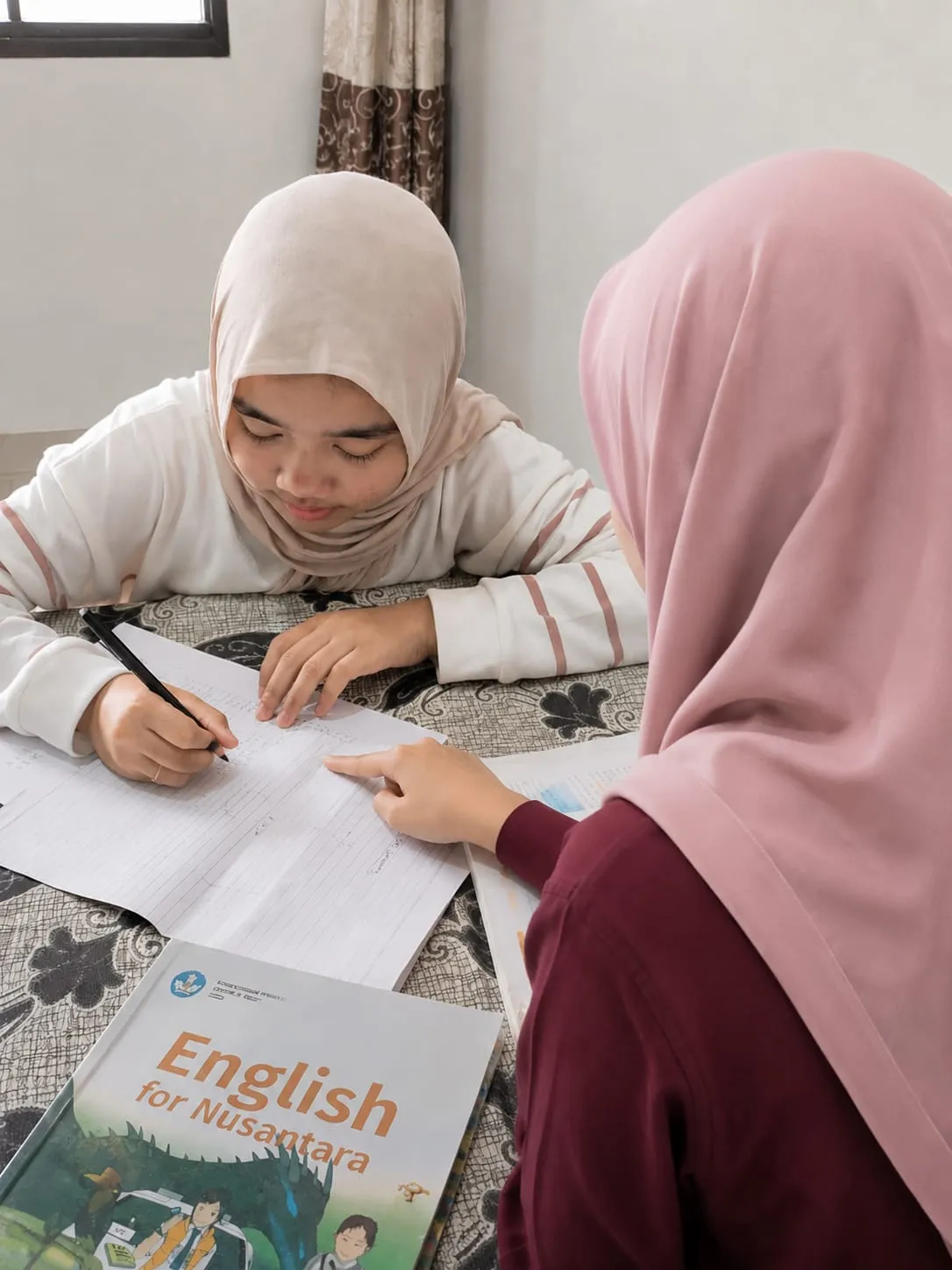 Seorang tutor berhijab mendampingi satu siswi yang sedang menulis di buku tulis saat les di rumah, dengan buku pelajaran English for Nusantara di atas meja.
